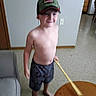 child, boy, smiling, baseball_cap, shirtless, shorts, standing, indoors, portrait, face, person, wooden_stick, coffee_table, living_room, couch, tile_floor, home, furniture, casual, happy