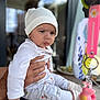 Dahlia Mila is registered to the contest to win money with this photo: baby, child, infant, white_clothing, hat, hand, table, keychain, pink_toy, indoor, portrait, person, cute, serious_expression, sitting, soft_lighting, blurred_background, adult_hand, cozy, casual