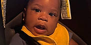 Damir is registered to the contest to win money with this photo: baby, infant, car_seat, yellow_bib, bracelet, dark_skin, curly_hair, hands, clothing, seat_belt, child, portrait, indoors, expression, cute, young_child, person, closeup, sitting, attention