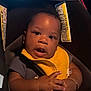 baby, infant, car_seat, yellow_bib, bracelet, dark_skin, curly_hair, hands, clothing, seat_belt, child, portrait, indoors, expression, cute, young_child, person, closeup, sitting, attention