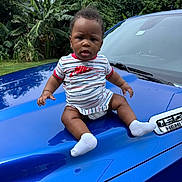 Damir is registered to the contest to win money with this photo: baby, child, blue, car_hood, truck, nike, striped_clothing, socks, bracelet, earrings, outdoor, banana_plants, greenery, nature, vehicle, portrait, curious, sitting, daylight, expression