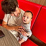 baby, child, young_man, striped_clothing, red_booth, wooden_wall, table, smile, curious, indoor, holding, person, seated, casual_clothing, portrait, tender_moment, family, human, happy, face