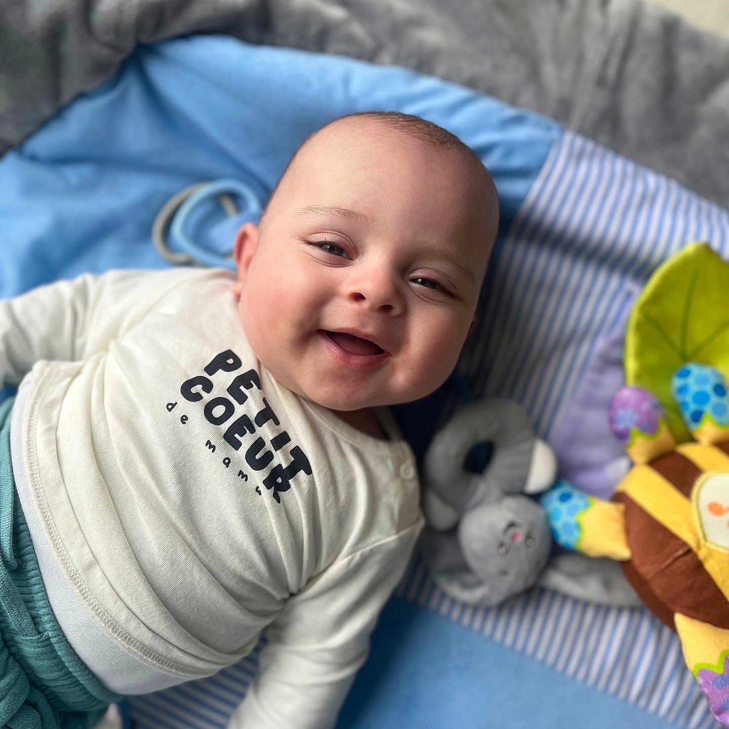 Lyam participe au concours pour gagner de l'argent avec cette photo : baby, blanket, child, clothing, colorful, cozy, cute, face, happy, indoor, infant, lying_down, person, playmat, portrait, shirt, smiling, soft_toy, toy, young_child