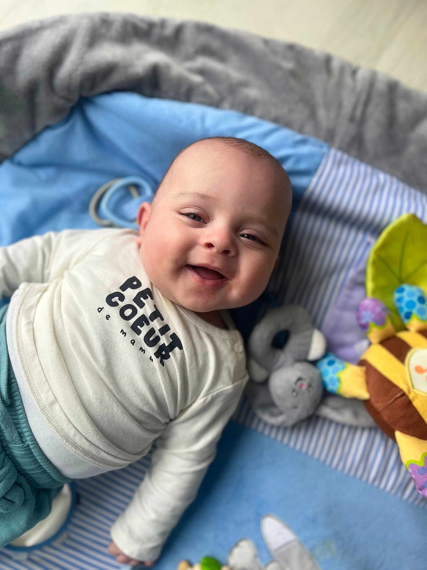 Lyam participe au concours pour gagner de l'argent avec cette photo : baby, blanket, child, clothing, colorful, cozy, cute, face, happy, indoor, infant, lying_down, person, playmat, portrait, shirt, smiling, soft_toy, toy, young_child