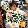 baby, child, car_seat, curly_hair, white_sweater, green_checkered_pants, white_shoes, blanket, indoor, carpet, chair, furniture, person, infant, cute, seated, curious, clothing, portrait, floor