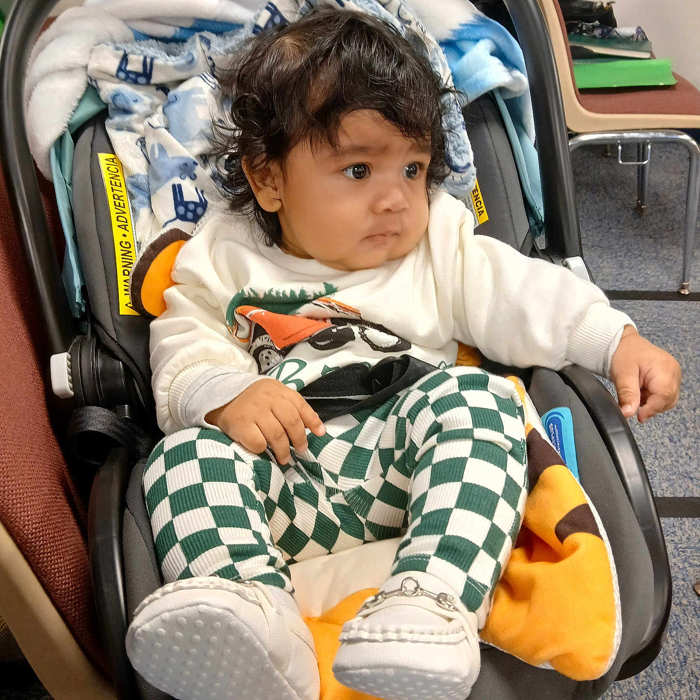 Emmanuel Caicedo joined the competition — help win amazing prizes! baby, child, car_seat, curly_hair, white_sweater, green_checkered_pants, white_shoes, blanket, indoor, carpet, chair, furniture, person, infant, cute, seated, curious, clothing, portrait, floor