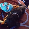Precious is registered to the contest to win money with this photo: person, dog, bed, blue_face_mask, indoor, brown_bedspread, black_dog, chain_collar, chewing_bone, lying_down, relaxation, casual, pet, companion, face, animal, comfort, home, resting, blur