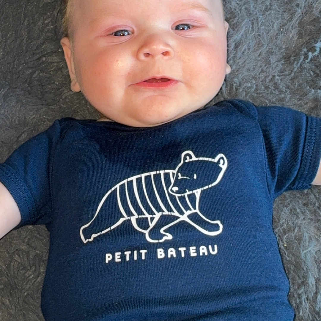 Ishaïm a rejoint le concours — aidez-le/la à gagner de superbes lots ! baby, bear_graphic, blue, child, chubby_cheeks, clothing, cute, face, fluffy_surface, gray_background, happy, head, infant, lying_down, onesie, petit_bateau, portrait, skin, smiling, text