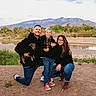 family, outdoor, river, mountains, dog, child, adult, casual_clothing, smiling, nature, grass, sand, water, trees, glasses, kneeling, happy, portrait, pets, people