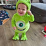 toddler, child, costume, green, monster, smiling, indoors, living_room, floor, wooden_floor, couch, pillow, play_mat, toy, happy, face, hood, feet, person, home