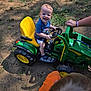 toddler, child, toy_tractor, outdoor, grass, dirt, smiling, steering_wheel, yellow_seat, green_toy, shorts, blue_shirt, footwear, person, happy, play, daylight, hand, head, nature