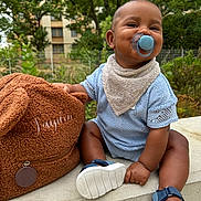Jayden a rejoint le concours — aidez-le/la à gagner de superbes lots ! baby, child, pacifier, blue_clothing, bib, sandals, bag, outdoor, greenery, building, concrete, happy, smiling, sitting, cute, infant, portrait, sunny, nature, daytime