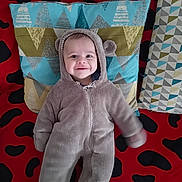 Idriss participe au concours pour gagner de l'argent avec cette photo : baby, child, onesie, hood, smile, pillow, pattern, red, black, blue, green, fabric, blanket, lying_down, cute, face, person, indoor, soft, cozy