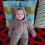 baby, child, onesie, hood, smile, pillow, pattern, red, black, blue, green, fabric, blanket, lying_down, cute, face, person, indoor, soft, cozy