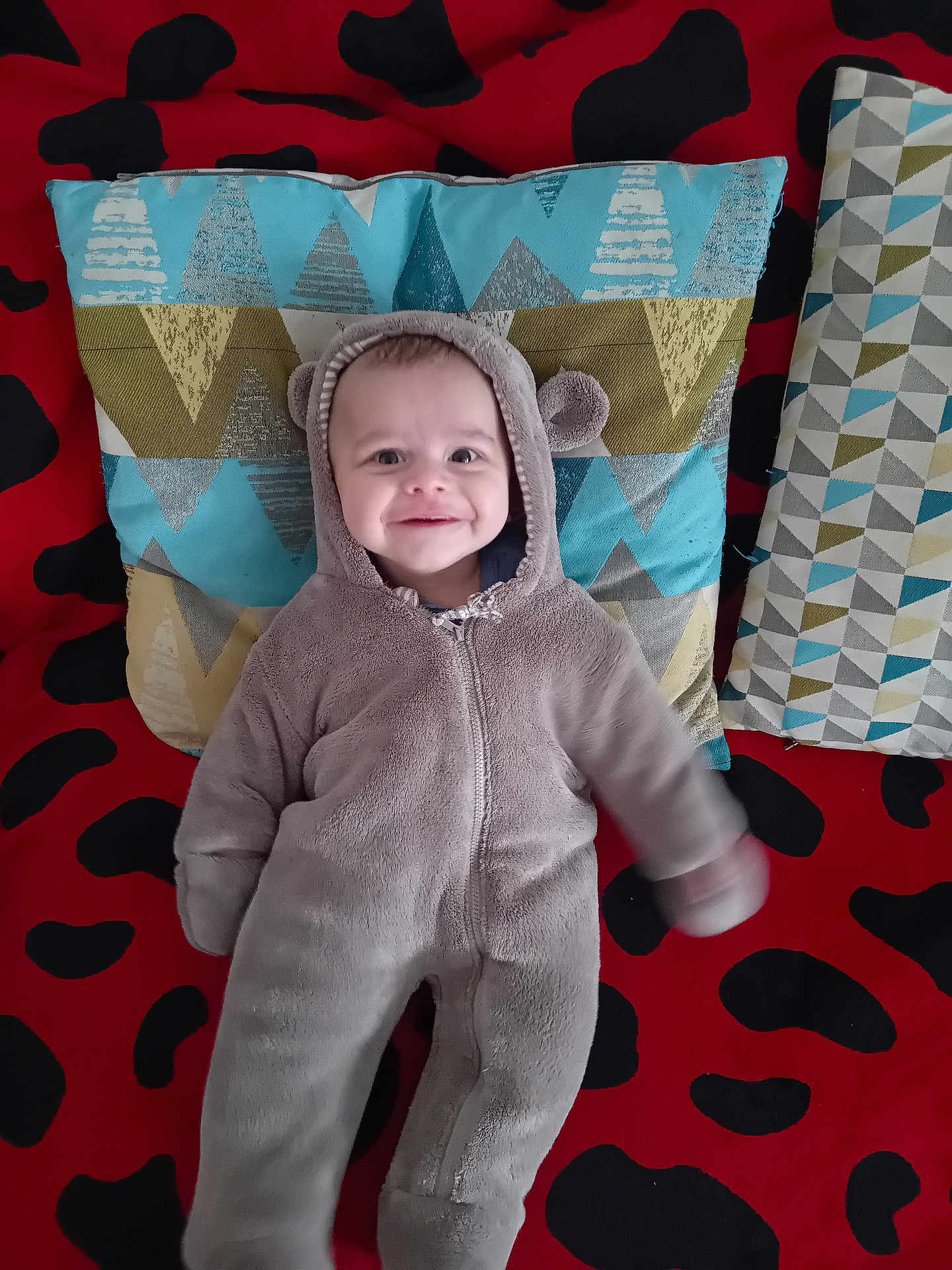 Idriss participe au concours pour gagner de l'argent avec cette photo : baby, child, onesie, hood, smile, pillow, pattern, red, black, blue, green, fabric, blanket, lying_down, cute, face, person, indoor, soft, cozy
