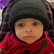 Maddix is registered to the contest to win money with this photo: baby, black_hat, casual_clothing, child, closeup, curious, face, hat, human, indoors, infant, knit_hat, looking_at_camera, person, plaid_pants, portrait, red_clothing, red_shirt, sitting, wide_eyes