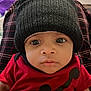 baby, black_hat, casual_clothing, child, closeup, curious, face, hat, human, indoors, infant, knit_hat, looking_at_camera, person, plaid_pants, portrait, red_clothing, red_shirt, sitting, wide_eyes