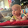 baby, child, colorful_clothing, red_blanket, indoor, living_room, curious, wide_eyes, face, person, furniture, chair, flag, window, curtain, household_items, floor, couch, hand, portrait