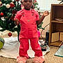 child, girl, toddler, pink_overalls, unicorn_slippers, christmas_tree, ornaments, braided_hair, hair_beads, window_blinds, bookshelf, carpet, living_room, holiday_decor, standing, portrait, indoors, neutral_expression, game_controller, pink_color