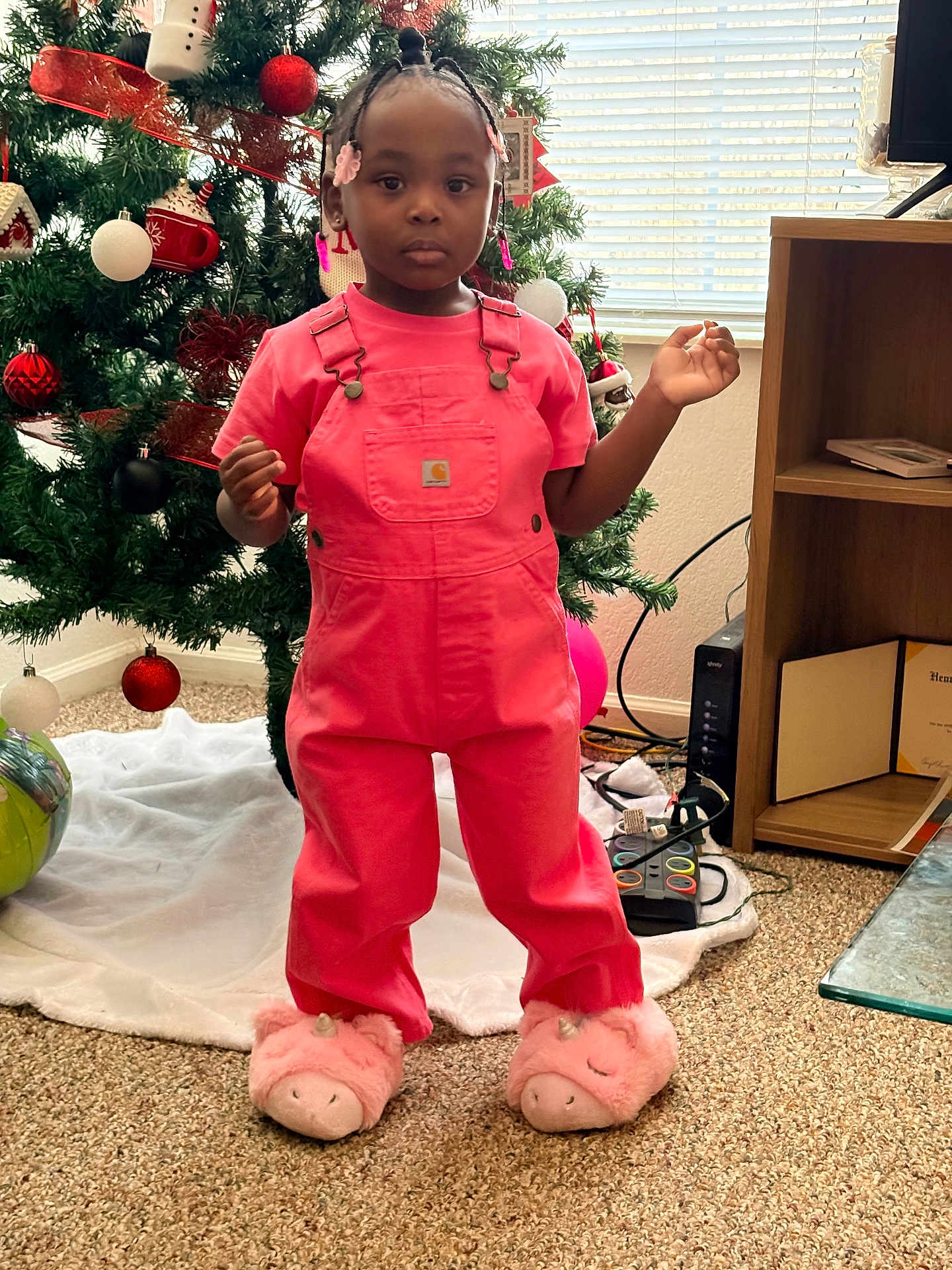 Autumn joined the competition — help win amazing prizes! child, girl, toddler, pink_overalls, unicorn_slippers, christmas_tree, ornaments, braided_hair, hair_beads, window_blinds, bookshelf, carpet, living_room, holiday_decor, standing, portrait, indoors, neutral_expression, game_controller, pink_color