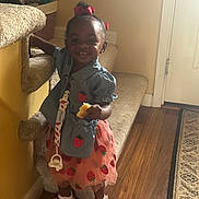 Paris is registered to the contest to win money with this photo: toddler, child, girl, smiling, indoor, carpet, staircase, handrail, dress, sandals, flooring, rug, snack, hair_bow, blue_top, pink_skirt, standing, holding, happy, person