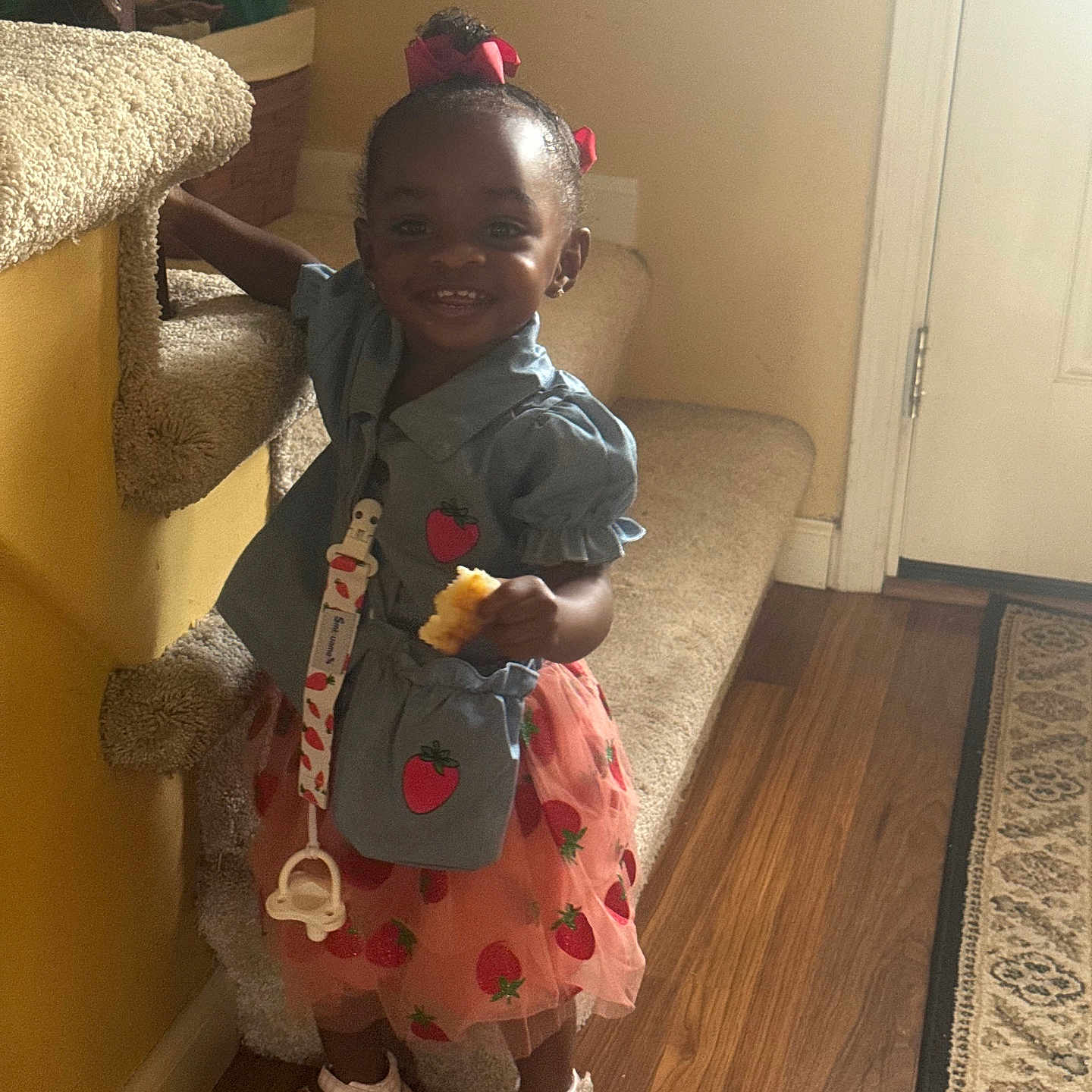 Paris is registered to the contest to win money with this photo: toddler, child, girl, smiling, indoor, carpet, staircase, handrail, dress, sandals, flooring, rug, snack, hair_bow, blue_top, pink_skirt, standing, holding, happy, person