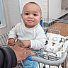 baby, child, infant, smiling, shopping_cart, supermarket, indoor, portrait, sitting, clothing, long_sleeves, jeans, socks, blanket, adult_hand, cart_handle, tile_wall, bathroom_sign, retail_store, happy