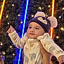 baby, child, infant, hat, bear_hat, onesie, hand, thumbs_up, neon_lights, fairy_lights, festive, background, person, face, warm_clothing, cute, portrait, indoor, celebration, holding