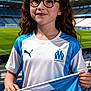 child, girl, glasses, curly_hair, stadium, flag, sports, soccer, team_jersey, blue_and_white, outdoor, smile, fan, young, happy, daytime, spectator, holding_flag, athletic, event