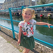 Victoire participe au concours pour gagner de l'argent avec cette photo : child, girl, smiling, blonde, pigtails, floral_top, green_skirt, sandals, river, railing, sidewalk, buildings, flowers, sunny, outdoor, urban, water, daylight, happy, posing