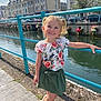 child, girl, smiling, blonde, pigtails, floral_top, green_skirt, sandals, river, railing, sidewalk, buildings, flowers, sunny, outdoor, urban, water, daylight, happy, posing