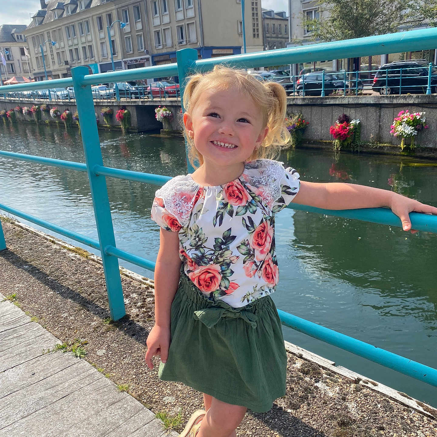 Victoire participe au concours pour gagner de l'argent avec cette photo : blonde, buildings, child, daylight, floral_top, flowers, girl, green_skirt, happy, outdoor, pigtails, posing, railing, river, sandals, sidewalk, smiling, sunny, urban, water