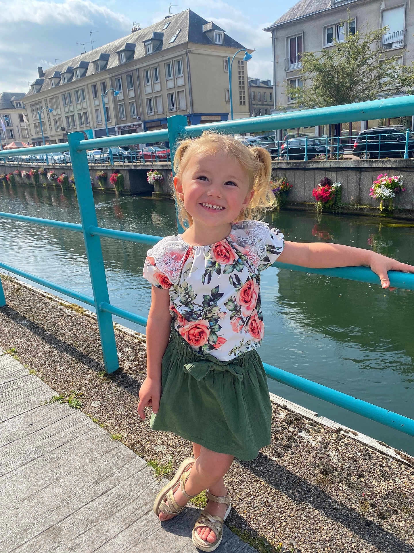 Victoire participe au concours pour gagner de l'argent avec cette photo : child, girl, smiling, blonde, pigtails, floral_top, green_skirt, sandals, river, railing, sidewalk, buildings, flowers, sunny, outdoor, urban, water, daylight, happy, posing
