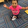 baby, child, smiling, pink_shirt, cross_legged, carpet, indoor, happy, person, cute, hair_puffs, toddler, colorful, striped_carpet, footwear, sitting, playroom, toy, floor, expression