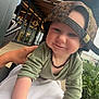 toddler, child, cap, leopard_print, necklace, tattoo, hand, outdoor, cafe, greenery, string_lights, smile, face, person, clothing, modern, building, window, daylight, casual