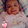 baby, infant, child, portrait, smiling, tongue_out, lying_down, pink_outfit, onesie, plaid_blanket, cushion, indoor, close_up, face, curly_hair, happy, heart_sticker, date_overlay, cartoon_print, photography