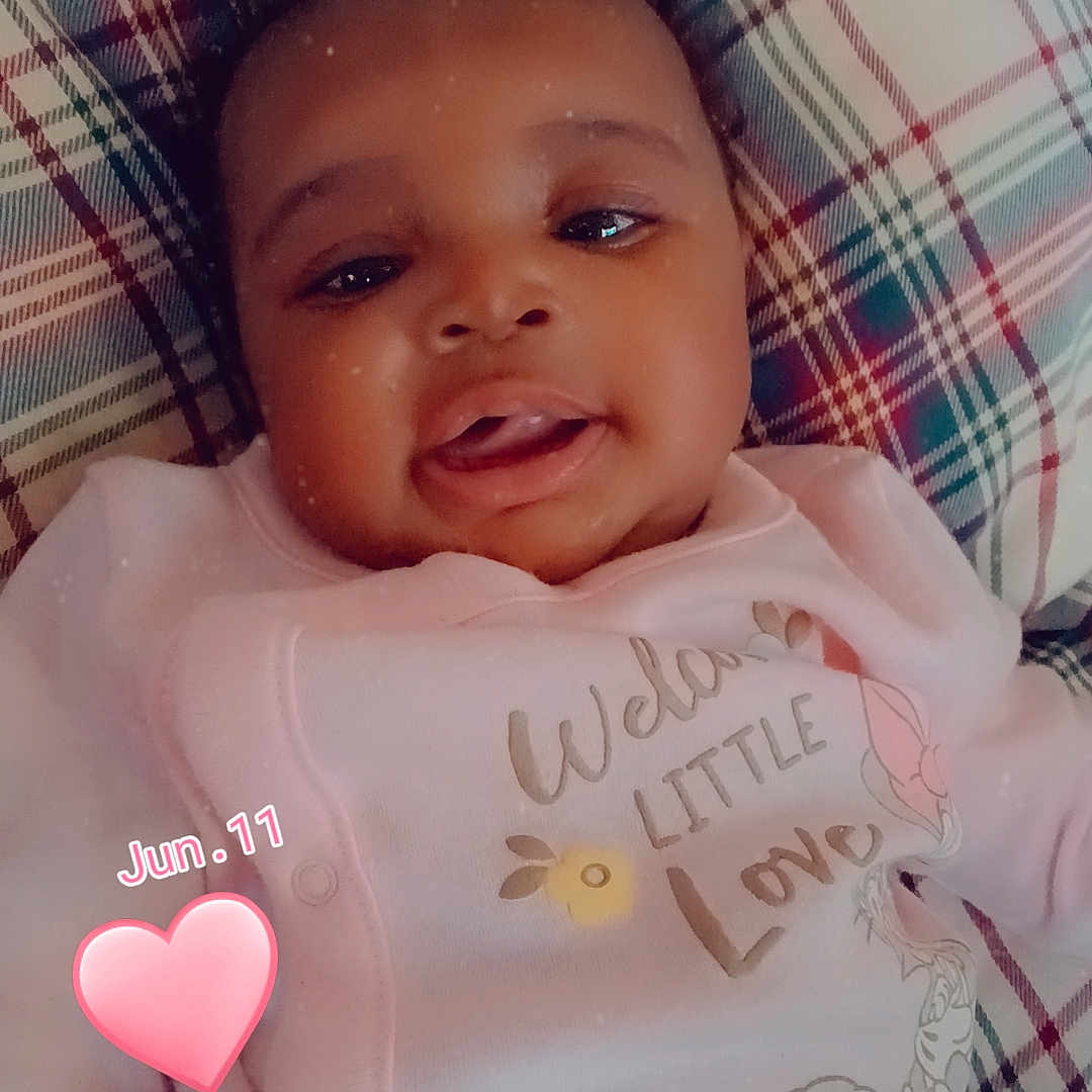 Ky'Manii is registered to the contest to win money with this photo: baby, infant, child, portrait, smiling, tongue_out, lying_down, pink_outfit, onesie, plaid_blanket, cushion, indoor, close_up, face, curly_hair, happy, heart_sticker, date_overlay, cartoon_print, photography