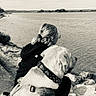 Bob a rejoint le concours — aidez-le/la à gagner de superbes lots ! dog, person, water, shoreline, lake, outdoors, nature, companion, sitting, harness, collar, leash, rocks, landscape, portrait, back_view, glasses, black_and_white, calm, friendship