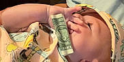Zaiden is registered to the contest to win money with this photo: baby, infant, child, dollar_bill, hand, face, hat, clothing, colorful, patterned, lying_down, indoors, cozy, curious, skin, person, young, cute, expression, holding