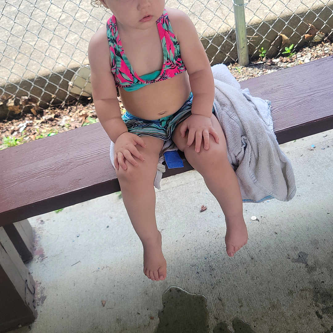 Dakota Gillam joined the competition — help win amazing prizes! toddler, child, swimsuit, bench, outdoor, fence, towel, water, concrete, curly_hair, summer, sitting, barefoot, expression, daylight, shade, play_area, young_child, casual, relaxed