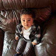 Isa is registered to the contest to win money with this photo: baby, child, infant, leather_couch, sitting, clothing, jacket, pants, socks, looking_up, curious, indoors, portrait, cute, person, young_child, cozy, casual_wear, home, seated