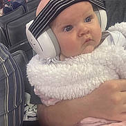 Lauren joined the competition — help win amazing prizes! baby, headphones, blanket, striped_headband, person, child, crowd, seated, indoor, event, clothing, holding, face, expression, pink, white, adult_hand, focus, noise_cancelling, ears