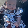 Lauren is registered to the contest to win money with this photo: baby, child, infant, person, holding, dress, patterned_clothing, outdoor, daylight, parking_lot, adult, hand, face, serious_expression, short_hair, palm_tree, sky, vehicle, shadow, sunlight