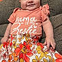 baby, child, smiling, sitting, couch, clothing, floral_skirt, peach_shirt, text_on_clothing, indoor, infant, cute, happy, person, portrait, furniture, skin, hand, face, head