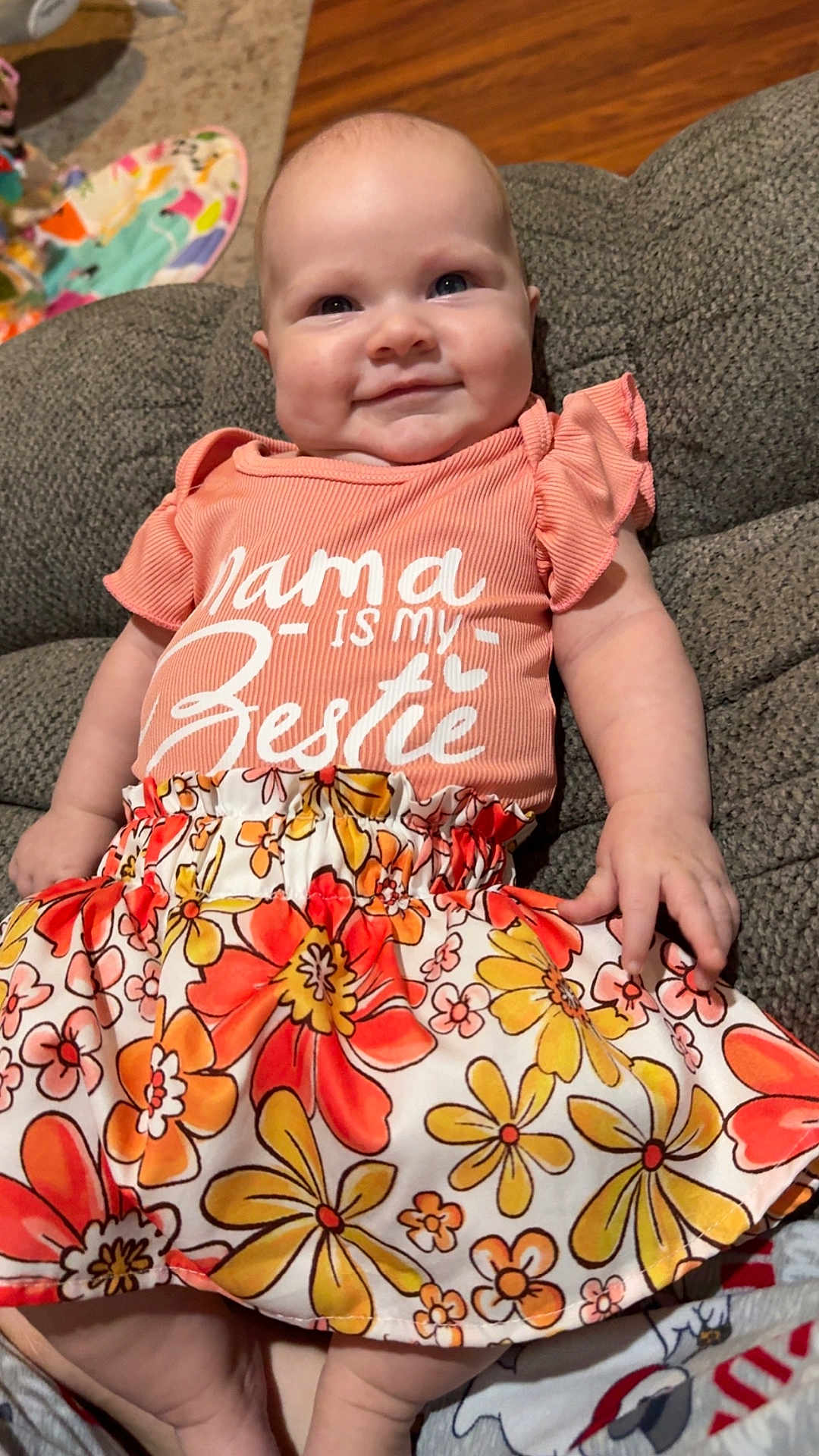 Lauren is registered to the contest to win money with this photo: baby, child, smiling, sitting, couch, clothing, floral_skirt, peach_shirt, text_on_clothing, indoor, infant, cute, happy, person, portrait, furniture, skin, hand, face, head