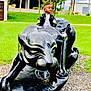 child, boy, sculpture, panther, black, jacket, outdoor, grass, greenery, statue, park, young, curious, portrait, casual_clothing, nature, daylight, person, looking_away, concrete