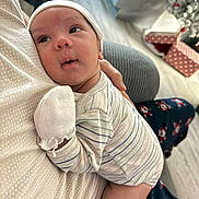 Jacob is registered to the contest to win money with this photo: baby, infant, striped_pajamas, mitten, resting, person, indoor, holiday_decor, gift_boxes, floor, comfort, closeup, headwear, lying_down, human, cute, peaceful, home, soft_lighting, cozy