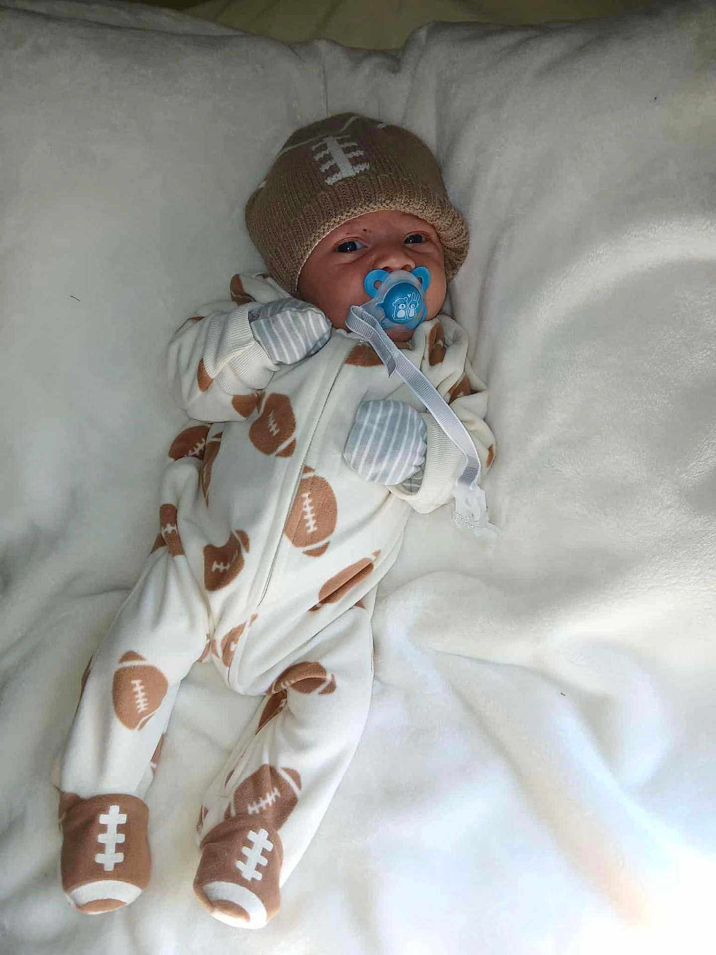 Kingston is registered to the contest to win money with this photo: baby, infant, pacifier, onesie, hat, football_pattern, blanket, sleepwear, cute, lying_down, gloves, footwear, indoors, soft_texture, newborn, child, portrait, cozy, comfort, sleeping_area