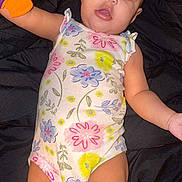 Aaliyah is registered to the contest to win money with this photo: baby, infant, child, onesie, floral_pattern, toy, colorful, blue_eyes, lying_down, curious, cute, skin, arm, legs, indoors, portrait, young_child, playful, expression, background