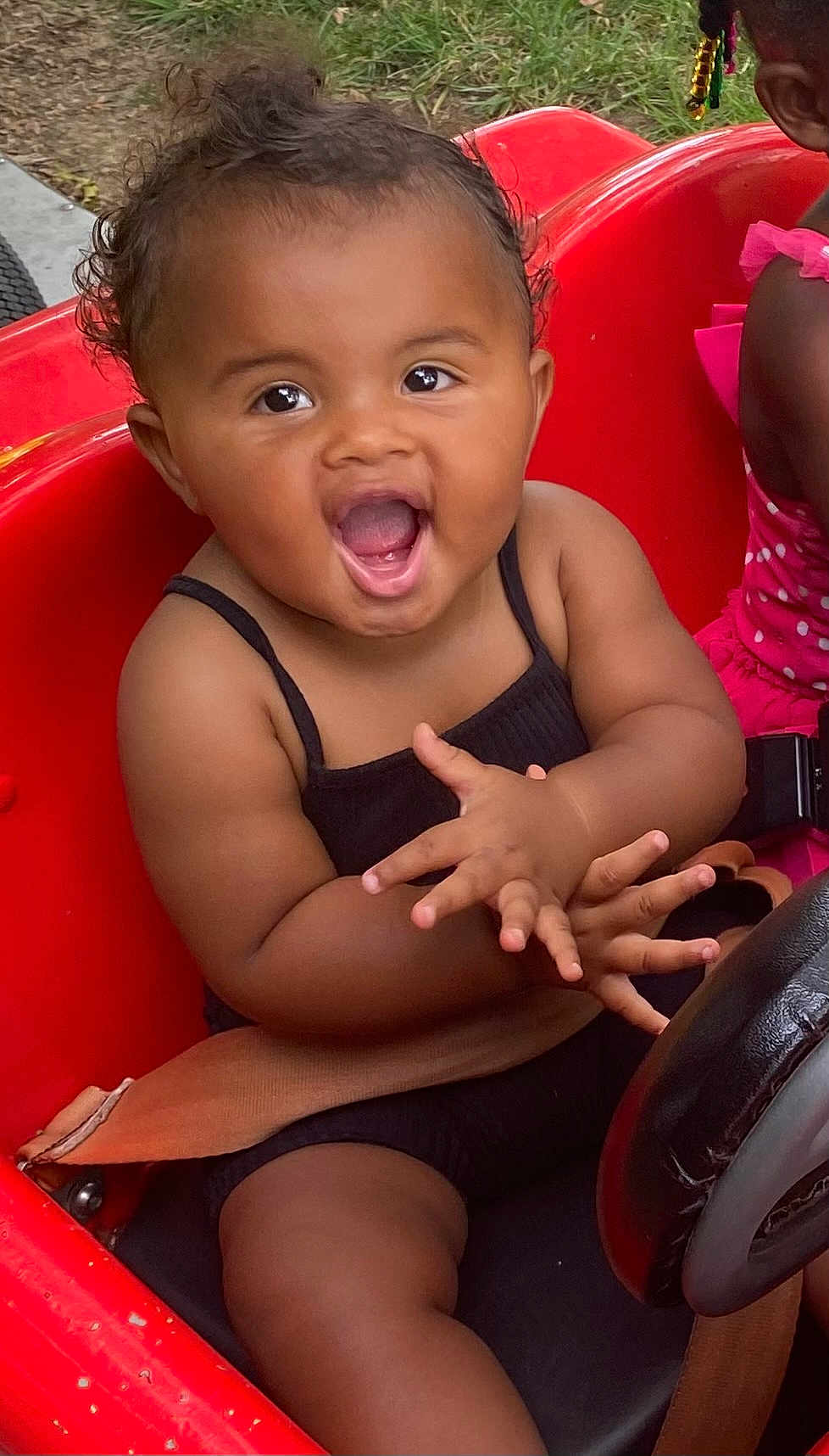 Ariel joined the competition — help win amazing prizes! toddler, child, smiling, clapping, curly_hair, black_outfit, red_toy_car, seatbelt, outdoor, grass, happy, person, driving_wheel, play, fun, cute, summer, sitting, joy, friend